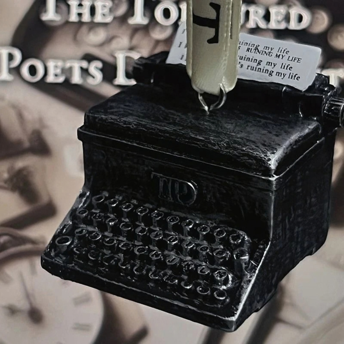 A black TTPD typewriter-shaped wall plaque or ornament with a small hanging tag, shown close-up against a sepia-toned background with blurred text and clock imagery.
