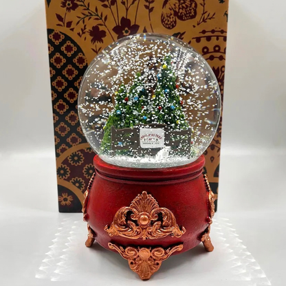 A red and gold ornate music box snow globe with a miniature decorated Christmas tree inside, set against a patterned gift box on a white surface.