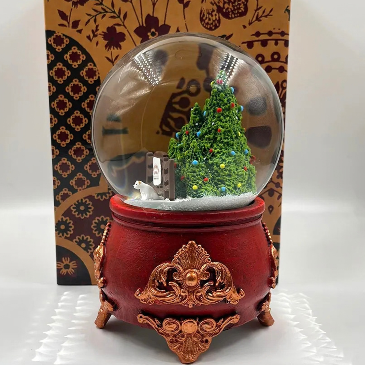 A Christmas-themed snow globe music box with a red ornate base, a clear glass globe containing a miniature decorated Christmas tree and white snow, set against a patterned brown and gold box.