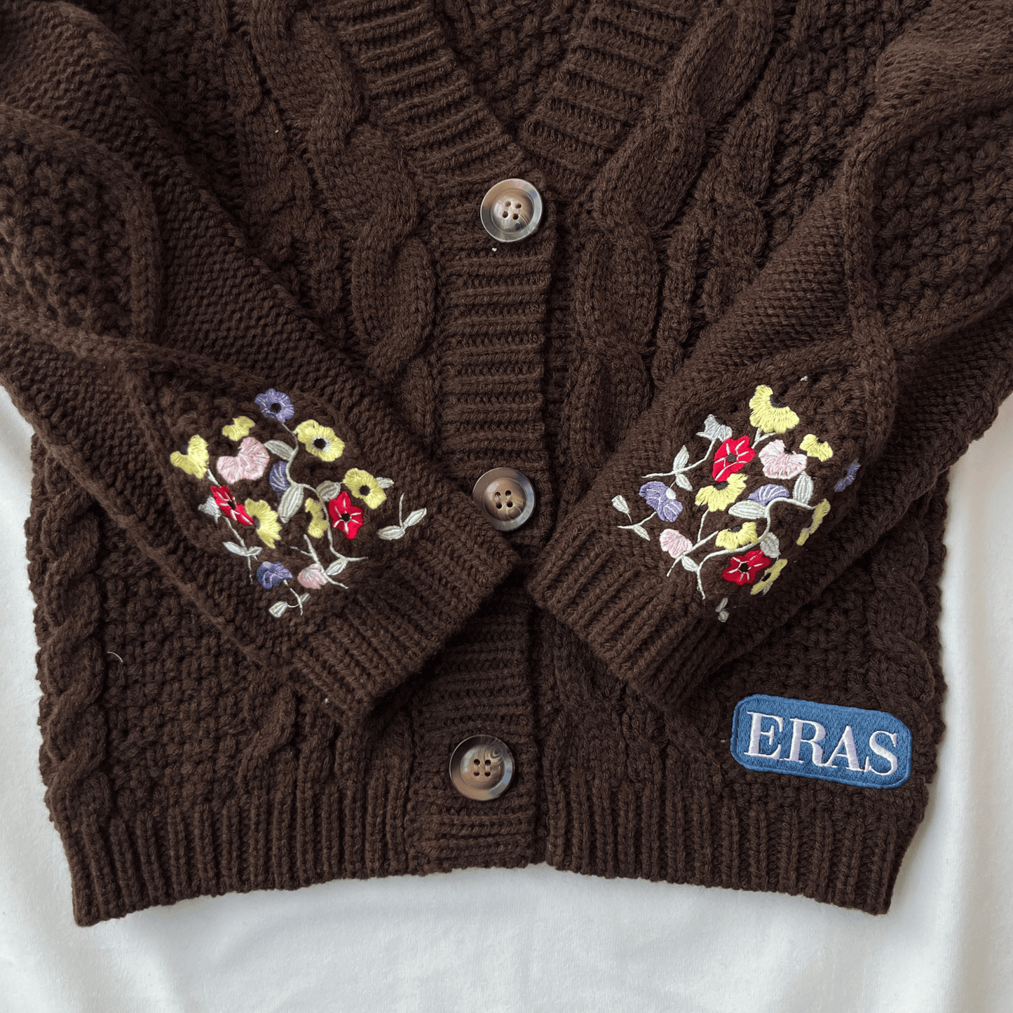 Brown cable knit cardigan with floral embroidery on sleeves and 'ERAS' logo on a white background