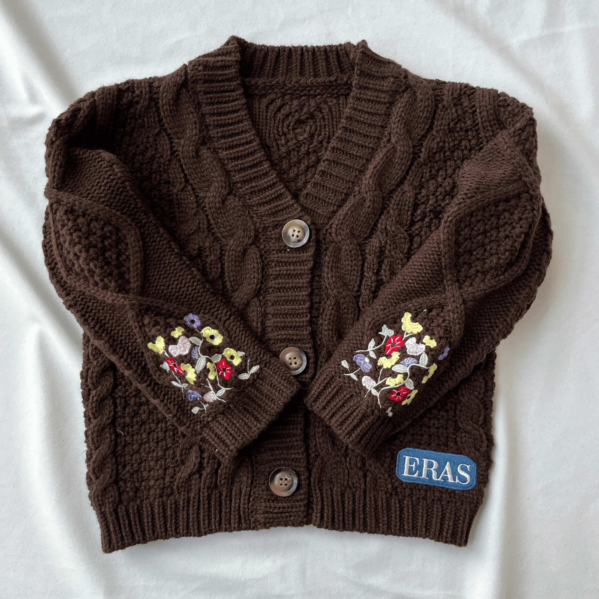 Brown knitted cardigan with floral embroidery on sleeves and 'ERAS' brand label, on a white background