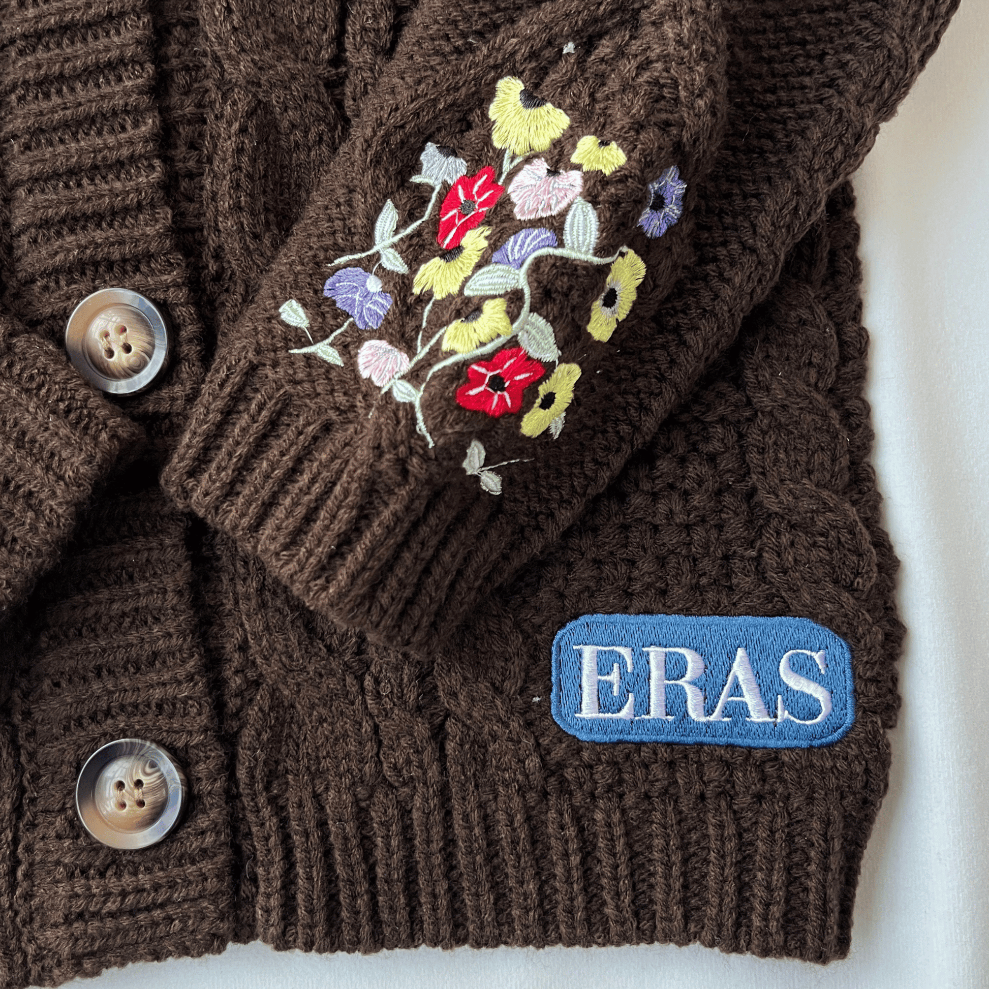 Brown knitted sweater with floral embroidery and 'ERAS' logo on a white background