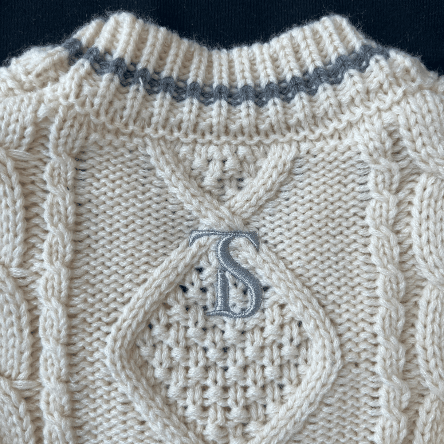 Close-up of a beige knitted sweater with a textured pattern and a monogram 'S' on a dark background.
