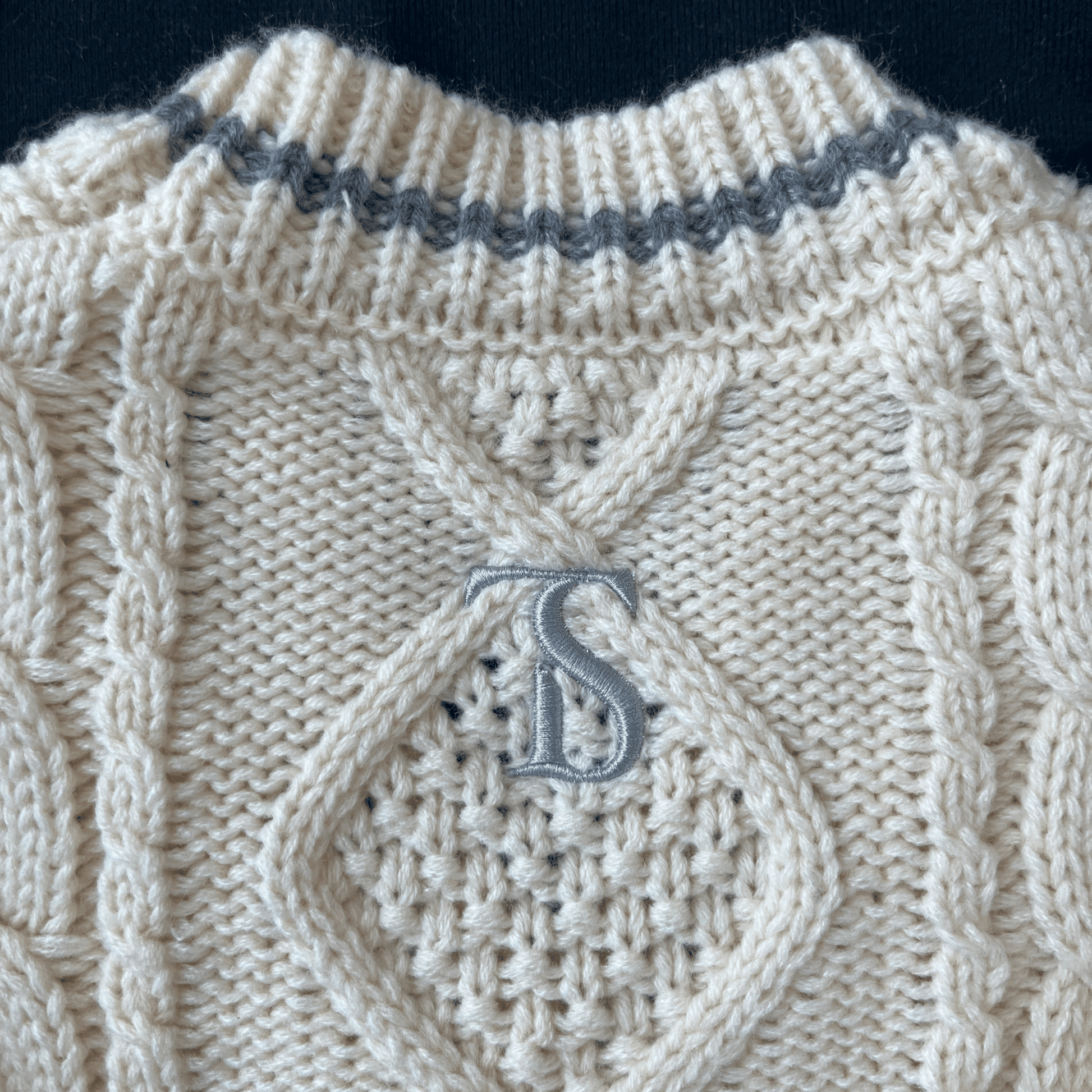 Close-up of a beige knitted sweater with a textured pattern and a monogram 'S' on a dark background.