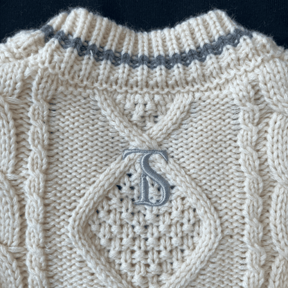 Close-up of a beige knitted sweater with a textured pattern and a monogram 'S' on a dark background.