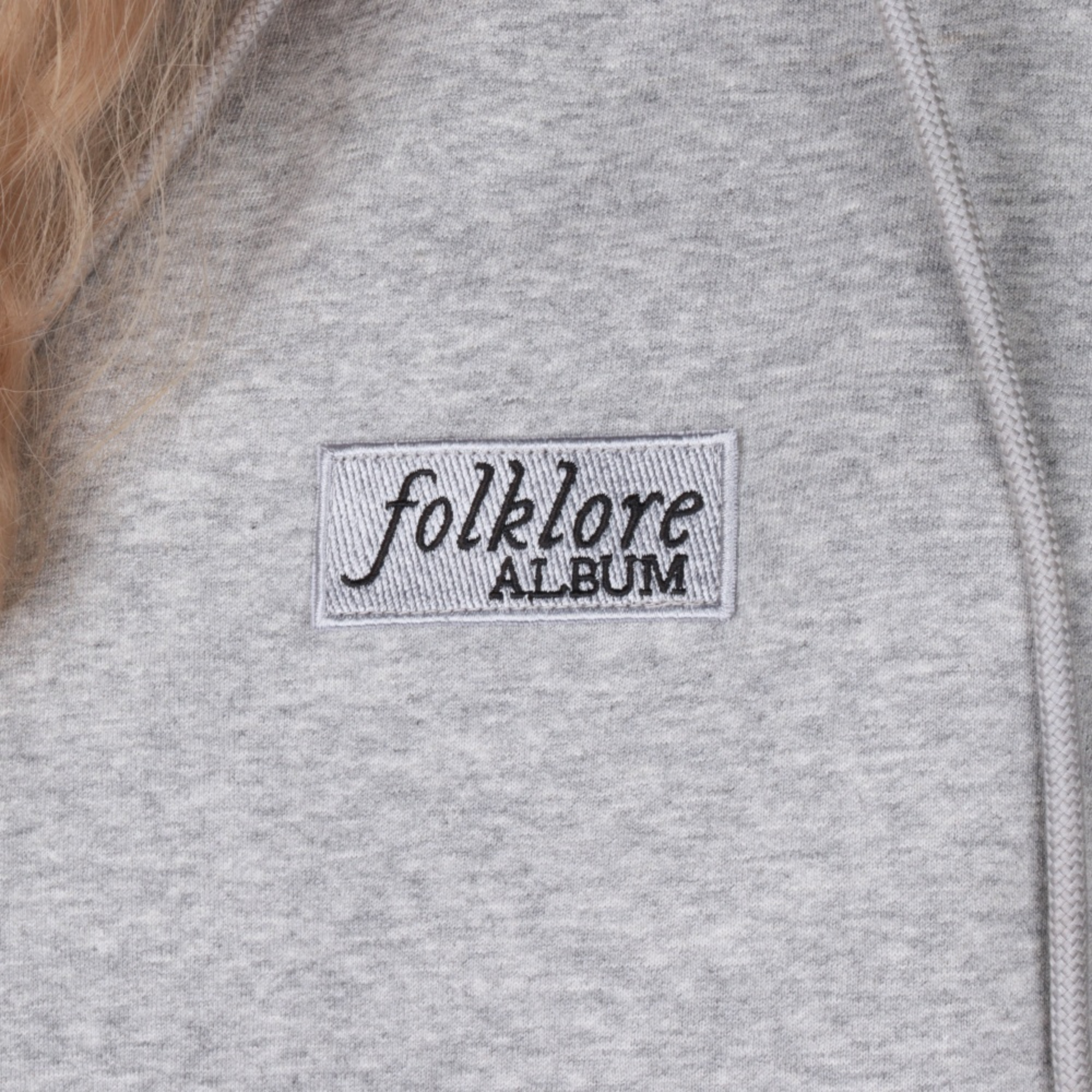 Close-up of a light gray heather zip-up hoodie featuring a rectangular embroidered patch on the chest with the text 'folklore ALBUM' in black.