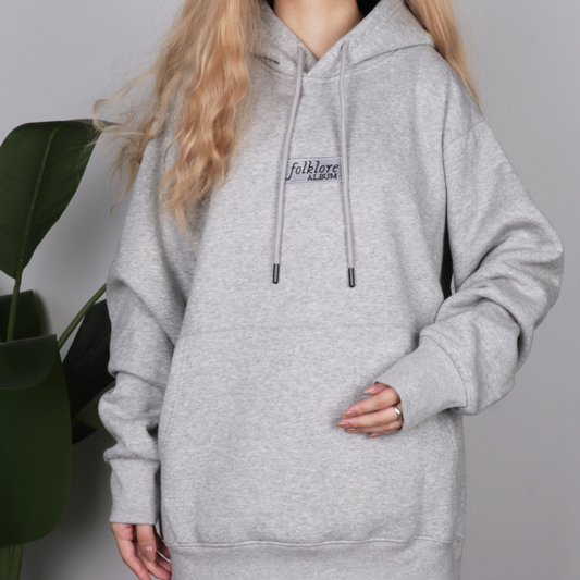 A person wearing an oversized light heather gray pullover hoodie with drawstrings and a small black 'folklore ALMOST' graphic on the chest, photographed against a plain light gray background with a green plant at the side.