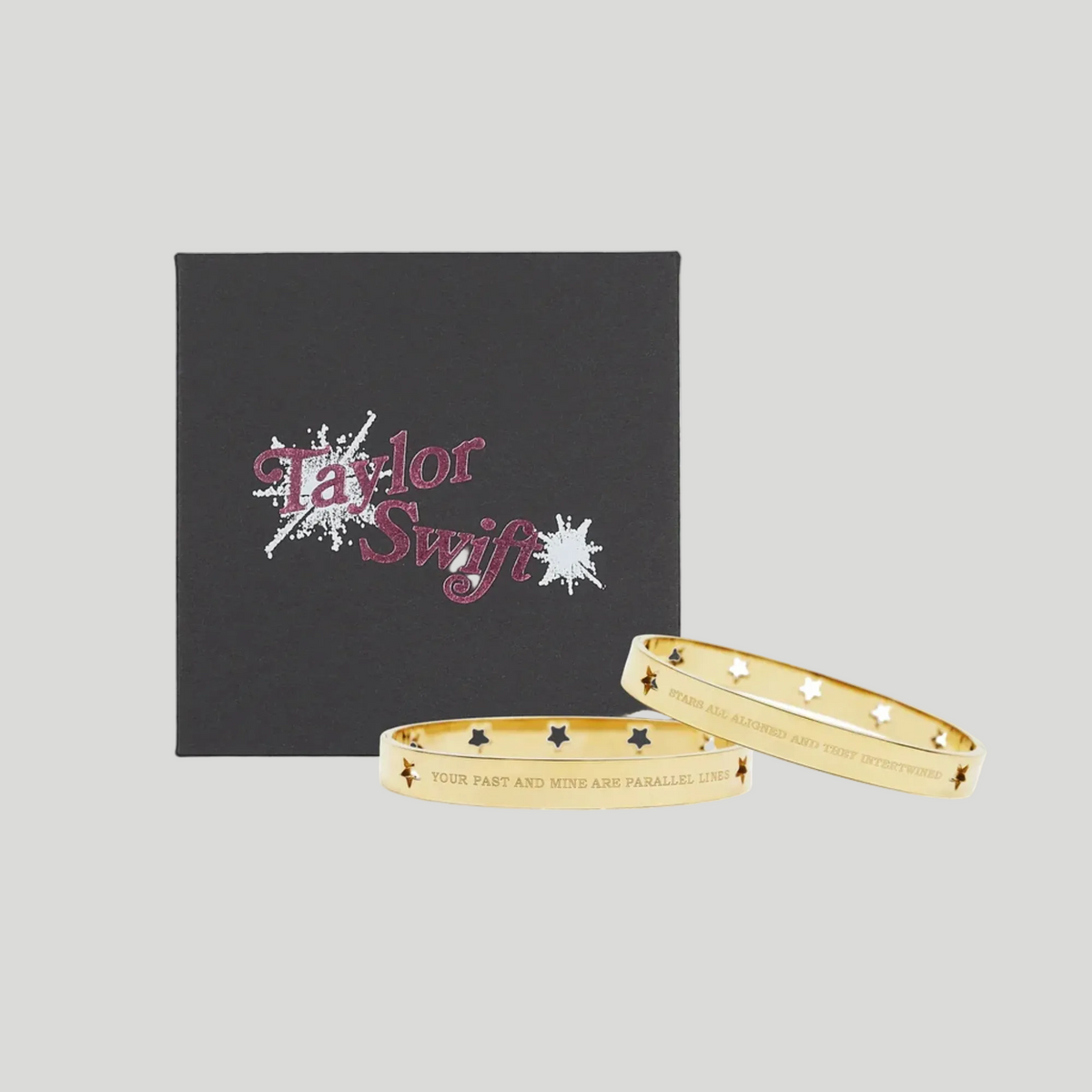Two gold bangle bracelets with star and heart details, displayed in front of a black Taylor Swift branded gift box on a white background.