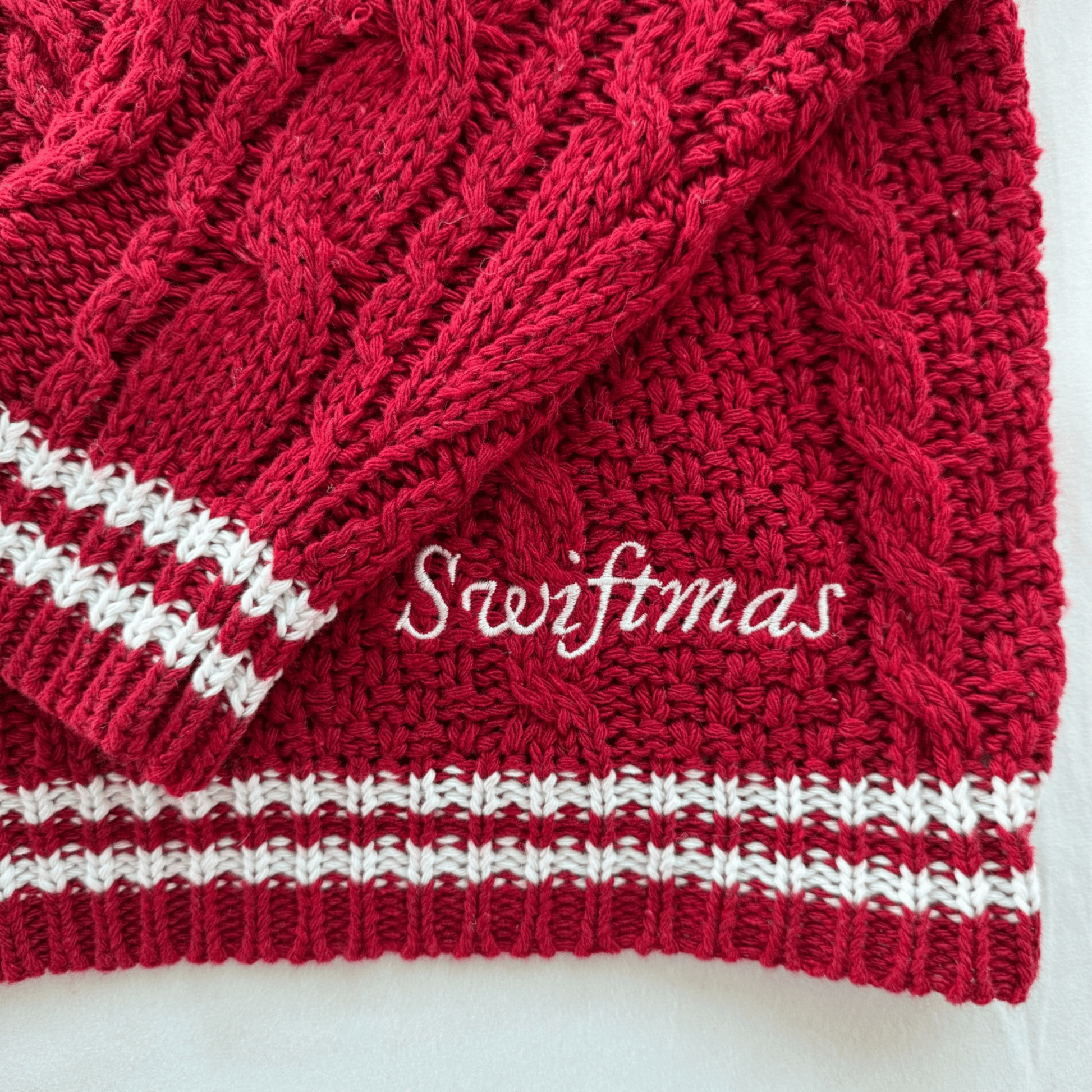 A close-up of a red cable-knit sweater or scarf with white striped ribbed trim and white cursive text reading 'Swiftmas' across the front, photographed on a light background.