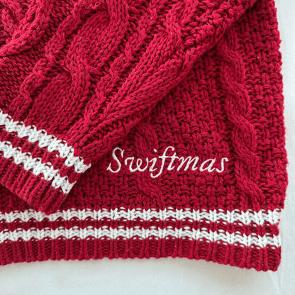 A close-up of a red cable-knit sweater or scarf with white striped ribbed trim and white cursive text reading 'Swiftmas' across the front, photographed on a light background.