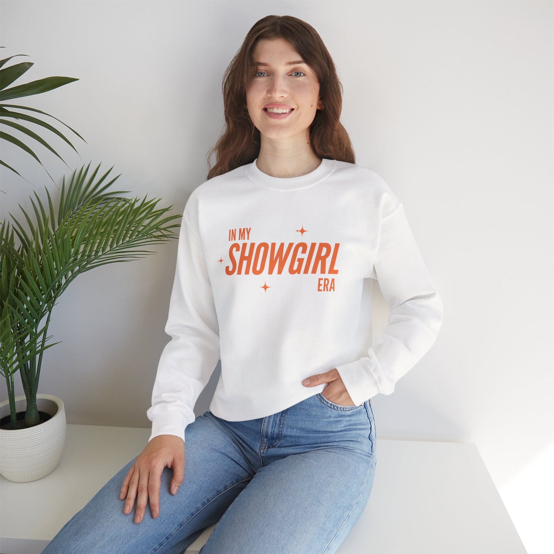 Woman wearing a white sweatshirt with 'In My Showgirl Era' text, sitting in a room with a plant.