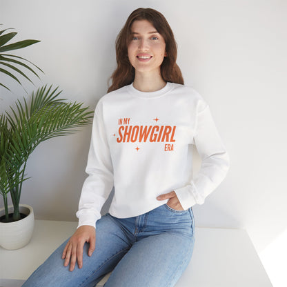 Woman wearing a white sweatshirt with 'In My Showgirl Era' text, sitting in a room with a plant.
