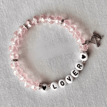 A pink faceted bead bracelet with white letter beads spelling 'LOVER' and heart-shaped beads, accented by silver-toned spacers and a silver clasp, displayed on a textured white fabric background.