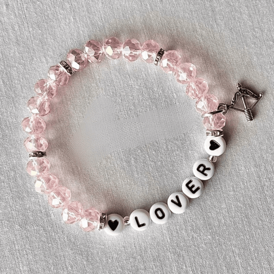 A pink faceted bead bracelet with white letter beads spelling 'LOVER' and heart-shaped beads, accented by silver-toned spacers and a silver clasp, displayed on a textured white fabric background.