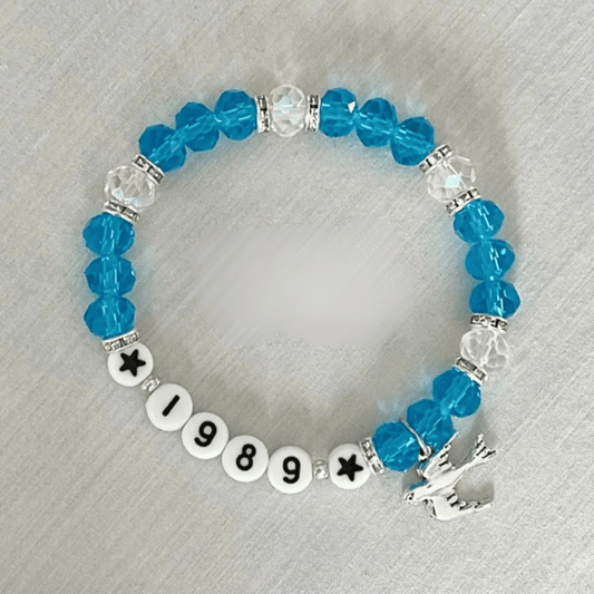 A beaded stretch bracelet featuring bright blue faceted beads, clear crystal spacer beads, white round beads spelling '1989' with black star and number details, and a silver-toned unicorn charm, displayed on a light gray textured fabric background.