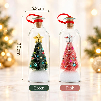 Two small Christmas tree glass bottle ornaments shown side by side on a marble surface with blurred holiday lights in the background. The left one is labeled Green and contains a miniature green Christmas tree with colorful ornaments and a gold star.