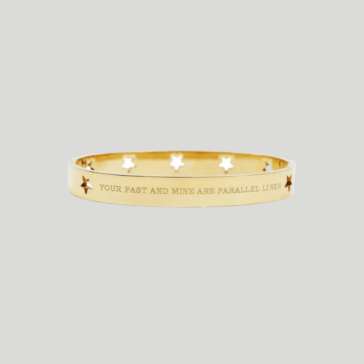 A gold bangle bracelet with star cutouts and the engraved text 'YOUR PAST AND MINE ARE PARALLEL LINES', shown on a plain white background.