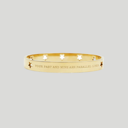 A gold bangle bracelet with star cutouts and the engraved text 'YOUR PAST AND MINE ARE PARALLEL LINES', shown on a plain white background.